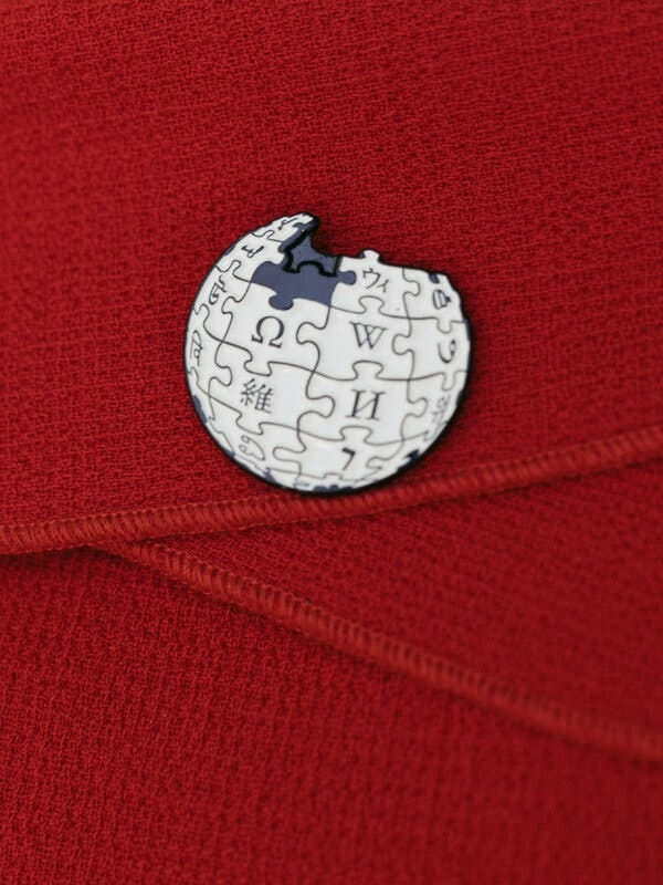 A round white pin decorated with shapes like puzzle pieces and the Wikipedia “W” on a red fabric background.
