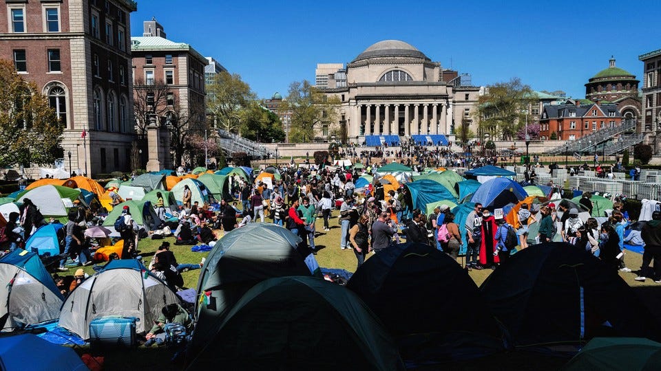 Pro-Palestinian protesters sit and stand, some in front of tents, in a large grassy area at Columbia University.
