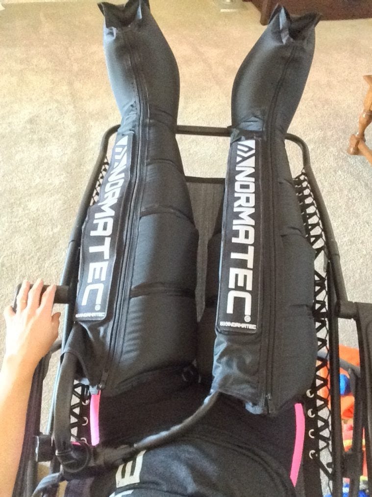 A person from waist down laying in a recliner with black compression boots from their feet ot upper thighs.White lettering spells out NORMOTEC on each boot A person from waist down laying in a recliner with black compression boots from their feet ot upper thighs.White lettering spells out NORMOTEC on each boot