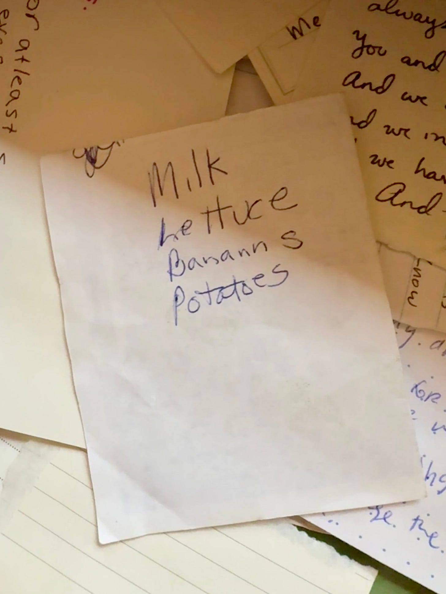 A scrap of paper, on top of other papers. It reads: Milk Lettuce Banann s Potatoes A scrap of paper, on top of other papers. It reads: Milk Lettuce Banann s Potatoes