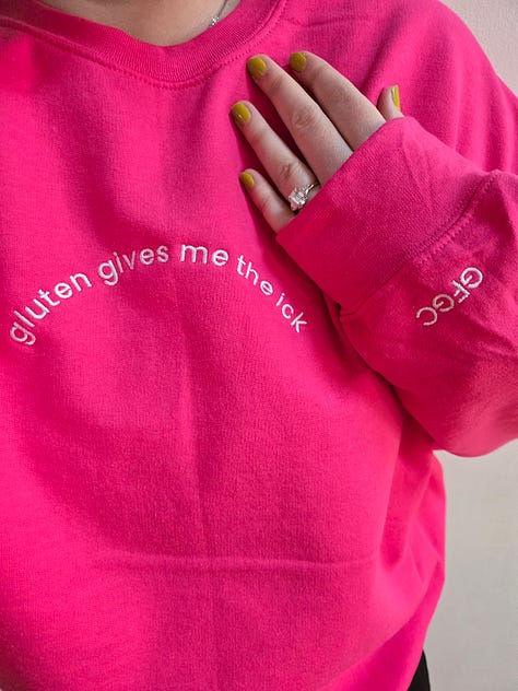 Modeling merch: A hot pink sweatshirt with white embroidered text on the front chest that says "gluten gives me the ick" and "GFGC" on the left sleeve near the wrist. A vintage wash denim baseball cap with an embroidered white pixelated chat bubble and black "GFGC" inside. A black sweatshirt with a green embroidered design on the front chest that says "GAME OVER, YOU ATE GLUTEN AND DIED" with a pixelated ox and wagon and "GFGC" on the left sleeve near the wrist.