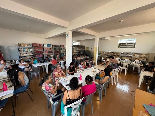 A imagem mostra um ambiente interno de biblioteca ou sala de leitura, com várias estantes repletas de livros ao fundo. O espaço é amplo e bem iluminado, com paredes brancas e piso de cerâmica marrom claro.  No ambiente, há cerca de vinte pessoas sentadas em cadeiras de plástico em torno de várias mesas brancas retangulares. A maioria são mulheres, algumas com crianças no colo. Elas parecem participar de uma atividade coletiva, possivelmente um encontro de estudo, leitura ou oficina. Algumas estão olhando livros ou cadernos, enquanto outras conversam entre si. Há garrafas de água e alguns materiais sobre as mesas.  O espaço é organizado, com as mesas distribuídas de forma a permitir circulação entre elas. A atmosfera transmite um clima de aprendizado colaborativo e troca de conhecimento, com atenção às atividades em andamento e interação entre os participantes.
