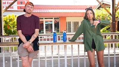 A man and a woman lean against a railing. She gestures a thumbs down.