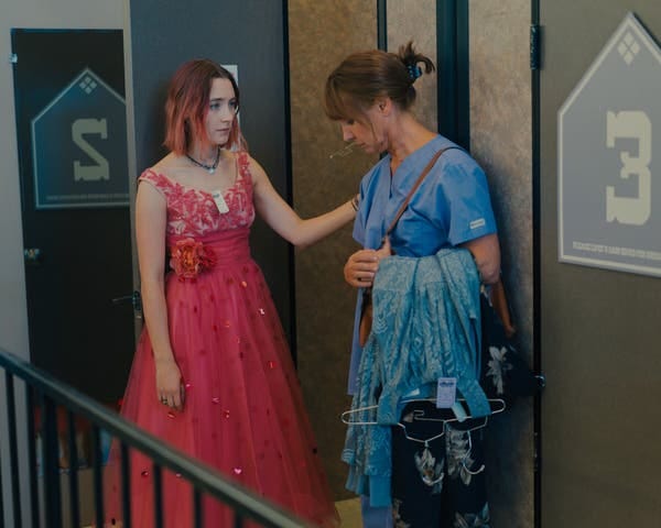 A young woman in a fancy hot pink dress rests a hand on an older woman in blue scrubs holding garments on hangers.