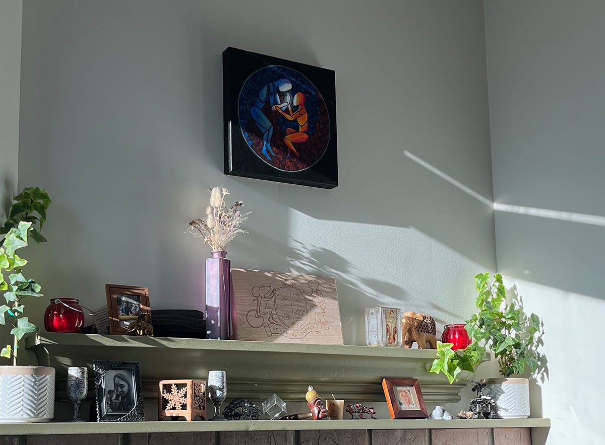 A painting of two stylized figures hangs over a fireplace. The paintings shows one figure of blue clasping hands with a fiery orange figure. From their joined hands pours a white light that illuminates them both.