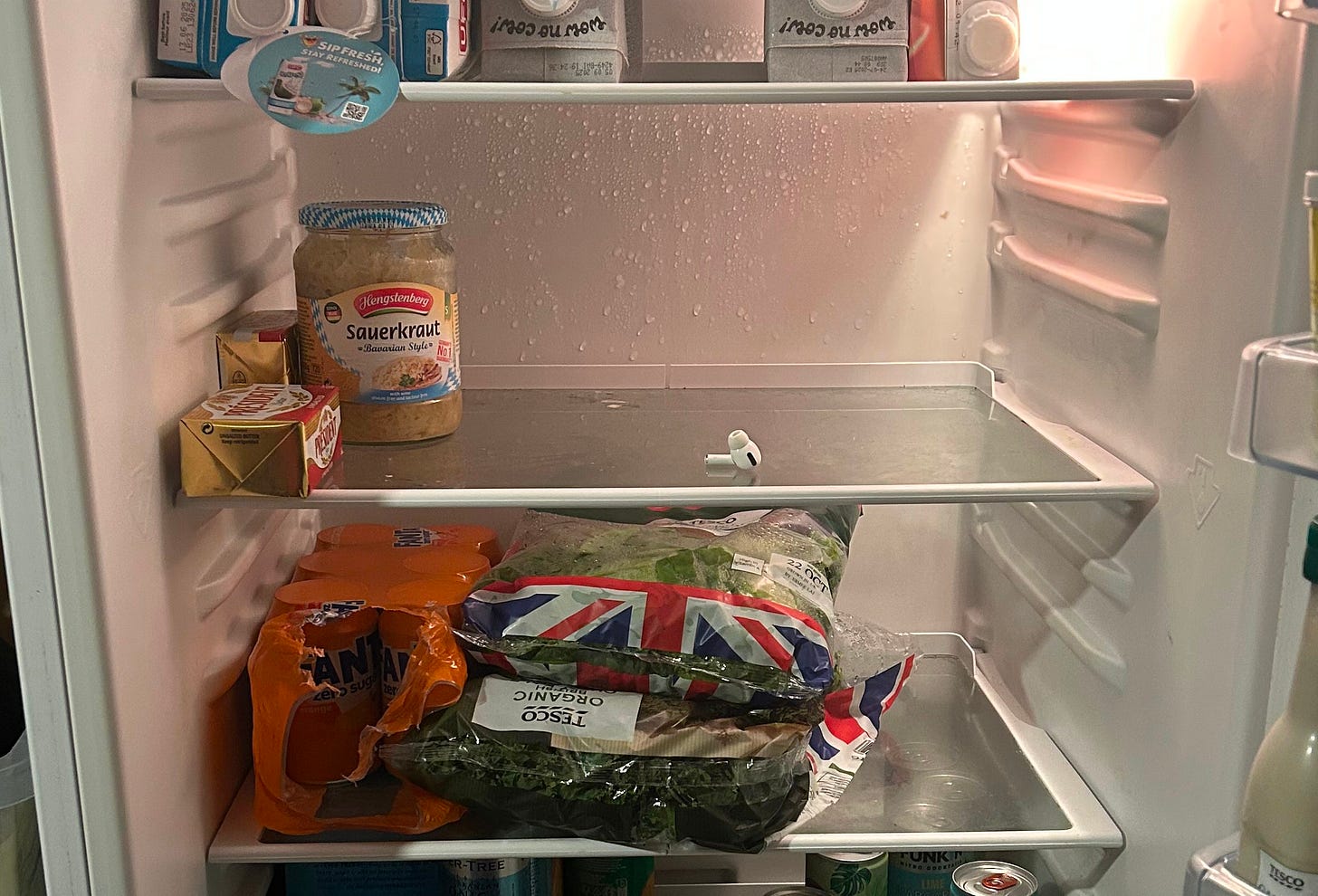 An interior shot of a fridge. There is a jar of sauerkraut, a pack of president unsalted butter and a single airpod on a shelf.