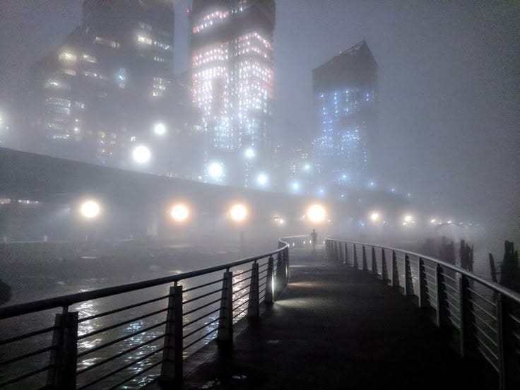This may contain: a foggy city street with lights on and buildings in the background