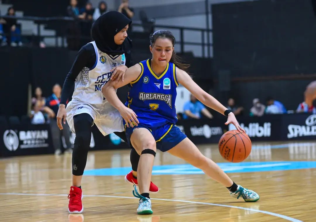An action shot from a Campus League Season 1 basketball game featuring two female student-athletes in a high-intensity play. In the foreground, a player from Universitas Airlangga in a blue and yellow jersey dribbles the ball with her right hand, leaning forward in a focused drive. She is being closely guarded by a player in a white and blue jersey and a black hijab, who has her hand extended to defend. The game is taking place on a polished indoor wooden court with a blurred crowd and digital sponsorship banners for "blibli" and "Spalding" in the background. The image captures the competitive spirit and athletic excellence that the new intercollegiate sports ecosystem aims to foster across Indonesia.