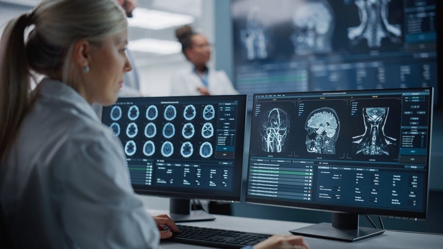 A medical professional analyzes brain and spine scans on two computer monitors in a clinical setting, while another person in a lab coat is blurred in the background. A medical professional analyzes brain and spine scans on two computer monitors in a clinical setting, while another person in a lab coat is blurred in the background.