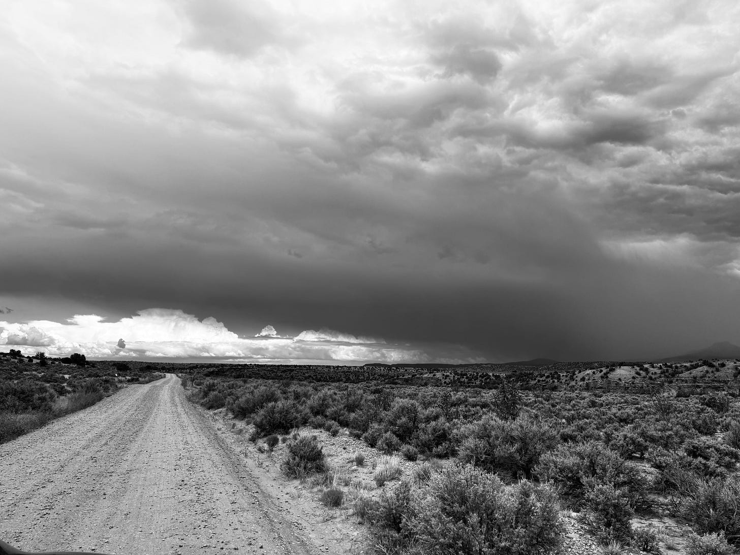 Black and white photo of a dirt road cutting through high desert scrubland near the Rio Chama in New Mexico, under a dramatic, heavily clouded sky with a distant storm cell visible on the horizon. No people or text are visible in the image.