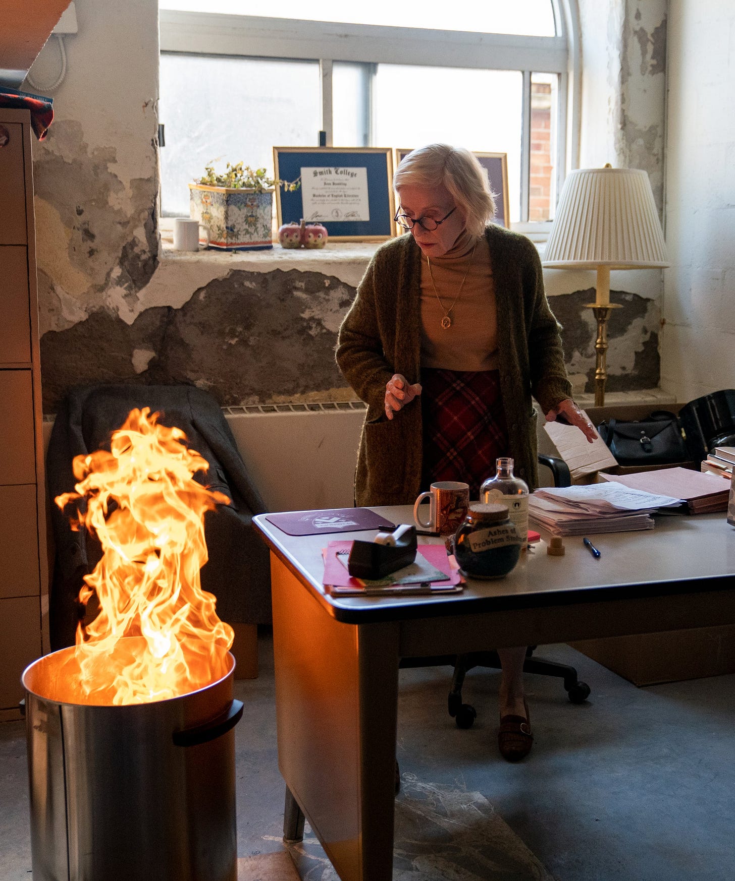 A veteran professor played by Holland Taylor in Netflix's The Chair in her basement office. Next to her desk is a trashcan that has a large flame coming out. The professor looks at the flame in alarm, realizing it's getting too big and might need to be put out.