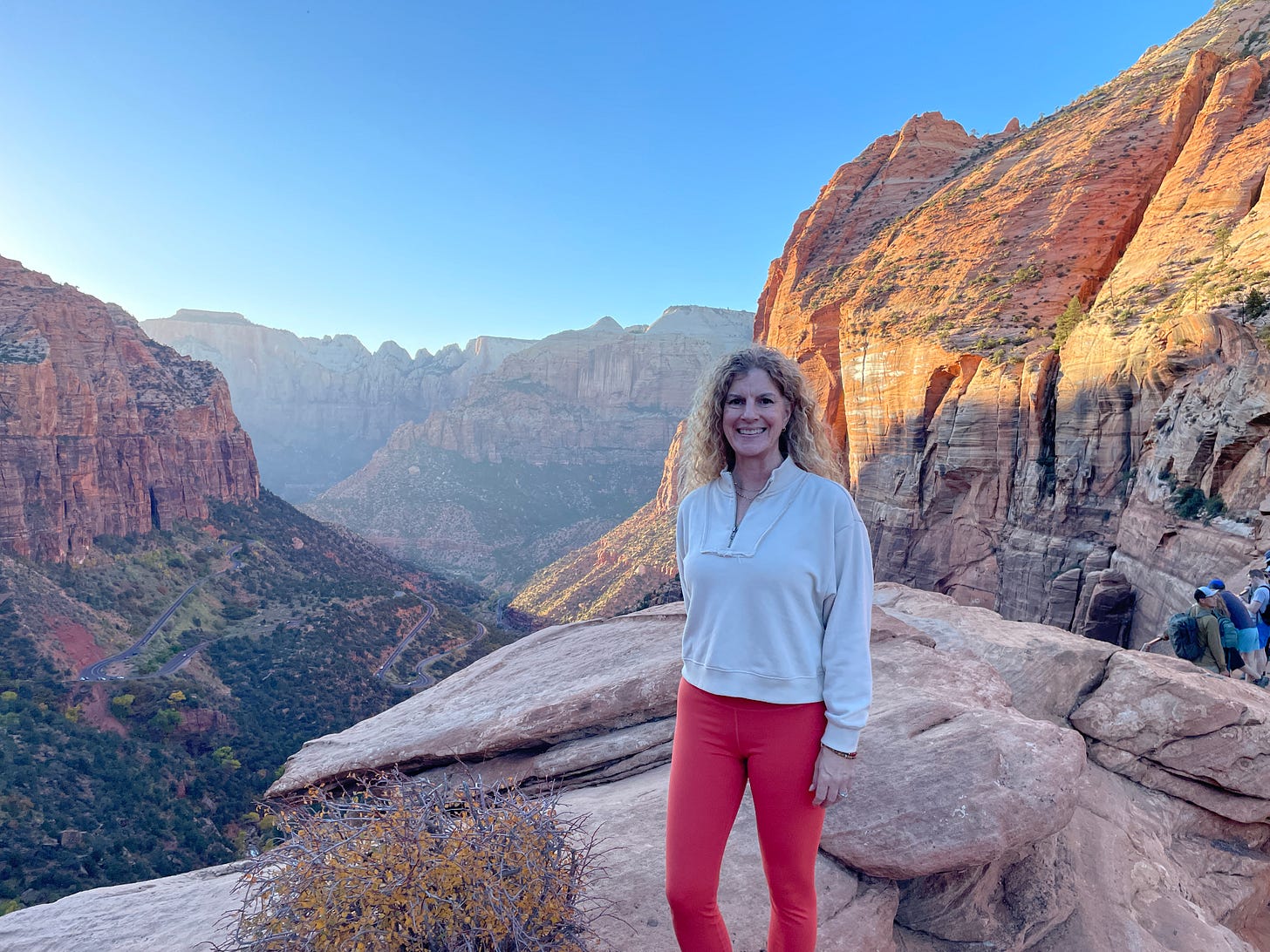Woman standing on a scenic overlook in Zion National Park with red rock canyon cliffs and winding road below at sunset.