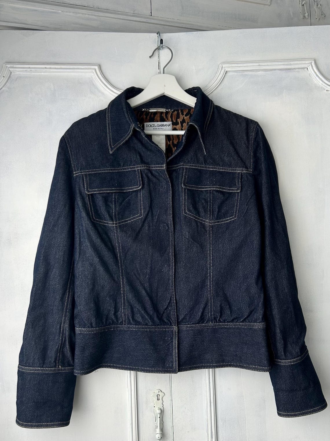 May include: A dark blue denim jacket with a collared neckline and two front pockets. The jacket has a fitted waist and long sleeves with cuffs. The interior lining of the collar features a leopard print. The jacket is hanging on a white hanger against a white background.