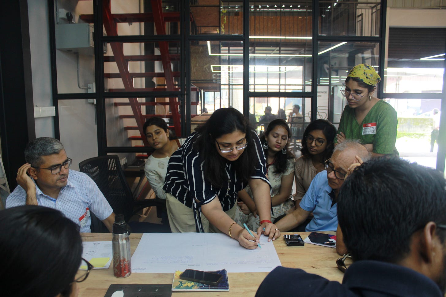 A group of people sitting around a table looking at a paper, where a lady is sketching something out to explain A group of people sitting around a table looking at a paper, where a lady is sketching something out to explain