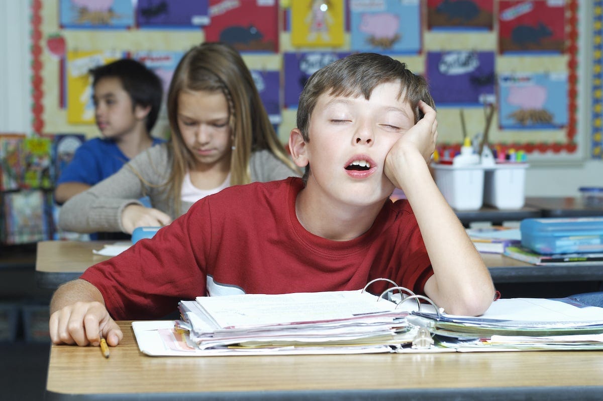 Boy in red shirt dozing at a school desk, head resting on his hand, atop a stack of binders; classmates blurred behind. Boy in red shirt dozing at a school desk, head resting on his hand, atop a stack of binders; classmates blurred behind.