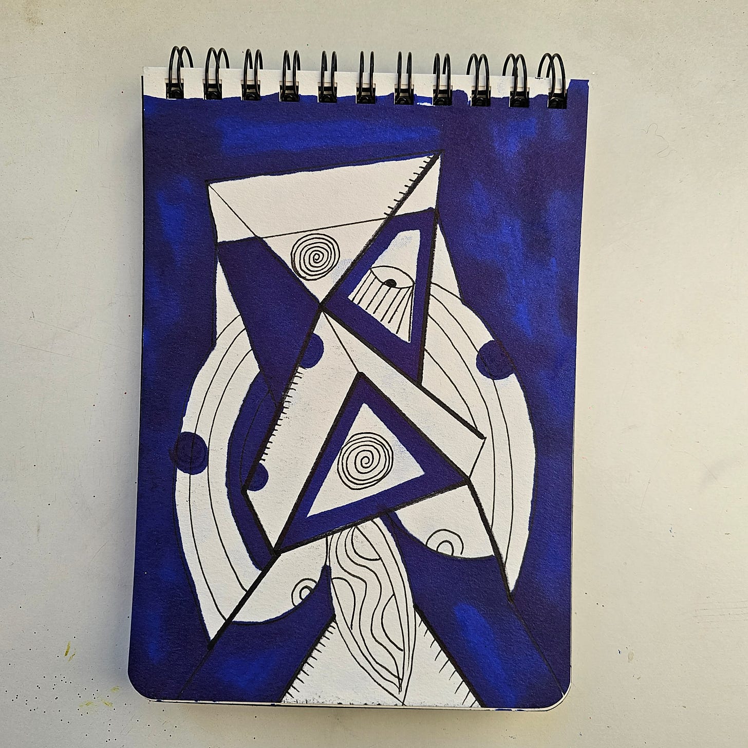 Alt Text:     A sketchbook drawing by artist FatbikeHero (b. 1983) featuring a central abstract geometric structure composed of triangles and circles. The design is filled with black ink patterns, including spirals and hatching, set against a solid, deep blue marker background. The style is raw and illustrative, reminiscent of neo-expressionism.