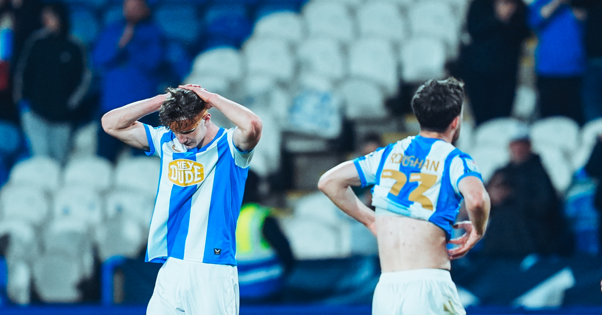 Try Huddersfield Town players hold their heads in their hands, with empty blue and white seats in the background. Try Huddersfield Town players hold their heads in their hands, with empty blue and white seats in the background.