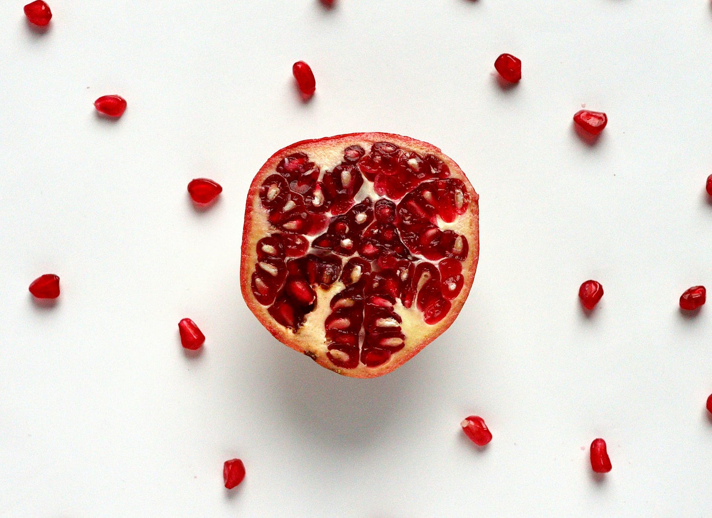 A pomegranate cut open lies at the center of a white background, its glistening red arils visible inside the fruit. Scattered around it, individual arils rest on the surface — some near, some farther out — as if the fruit and its seeds have spread across a wider field.