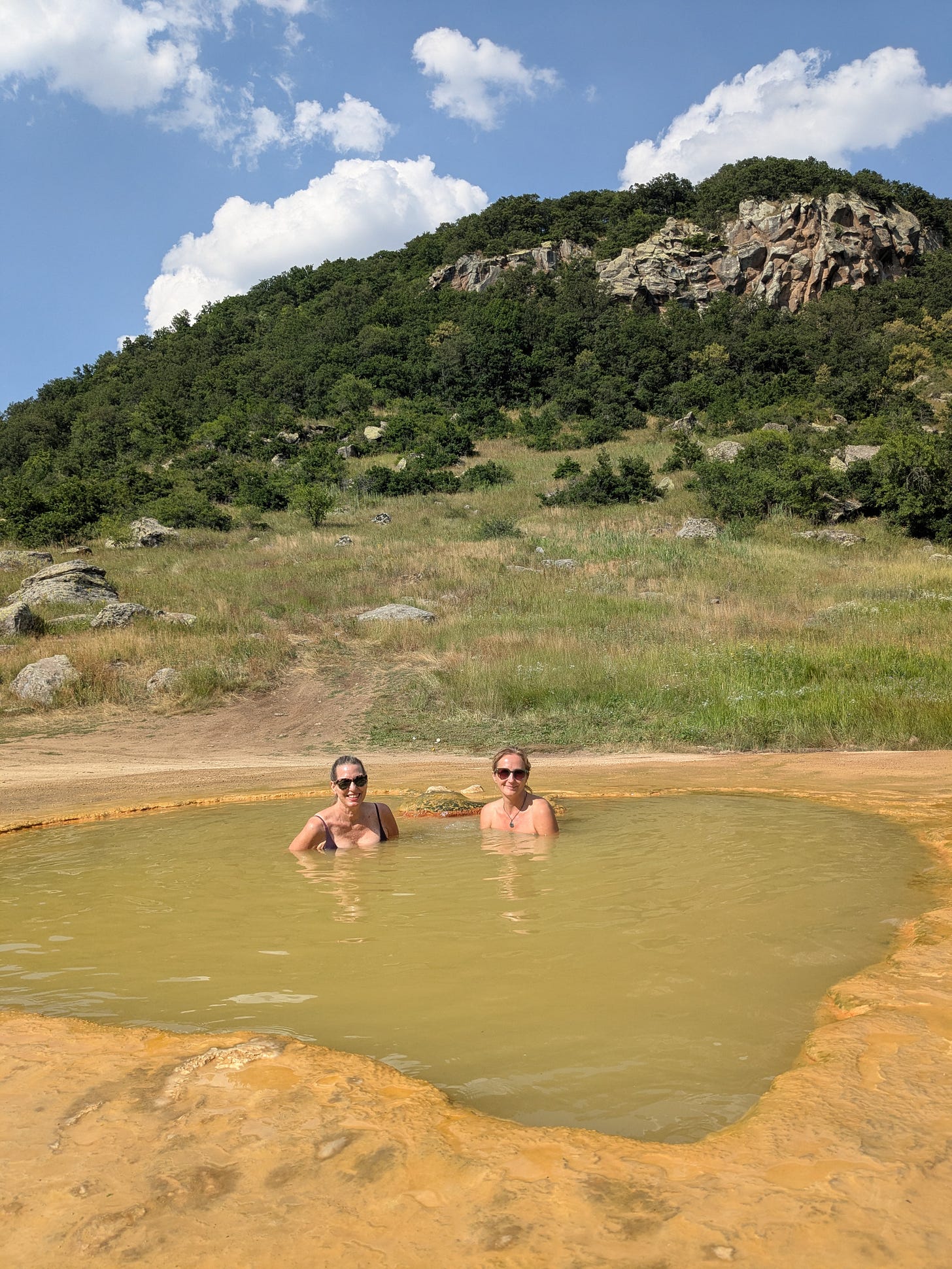 Two white women are smiling and wearing sunglasses with their hair pulled back. They are submerged in hot springs made of yellow clay. The mountain side and bright blue sky is behind them. Two white women are smiling and wearing sunglasses with their hair pulled back. They are submerged in hot springs made of yellow clay. The mountain side and bright blue sky is behind them.