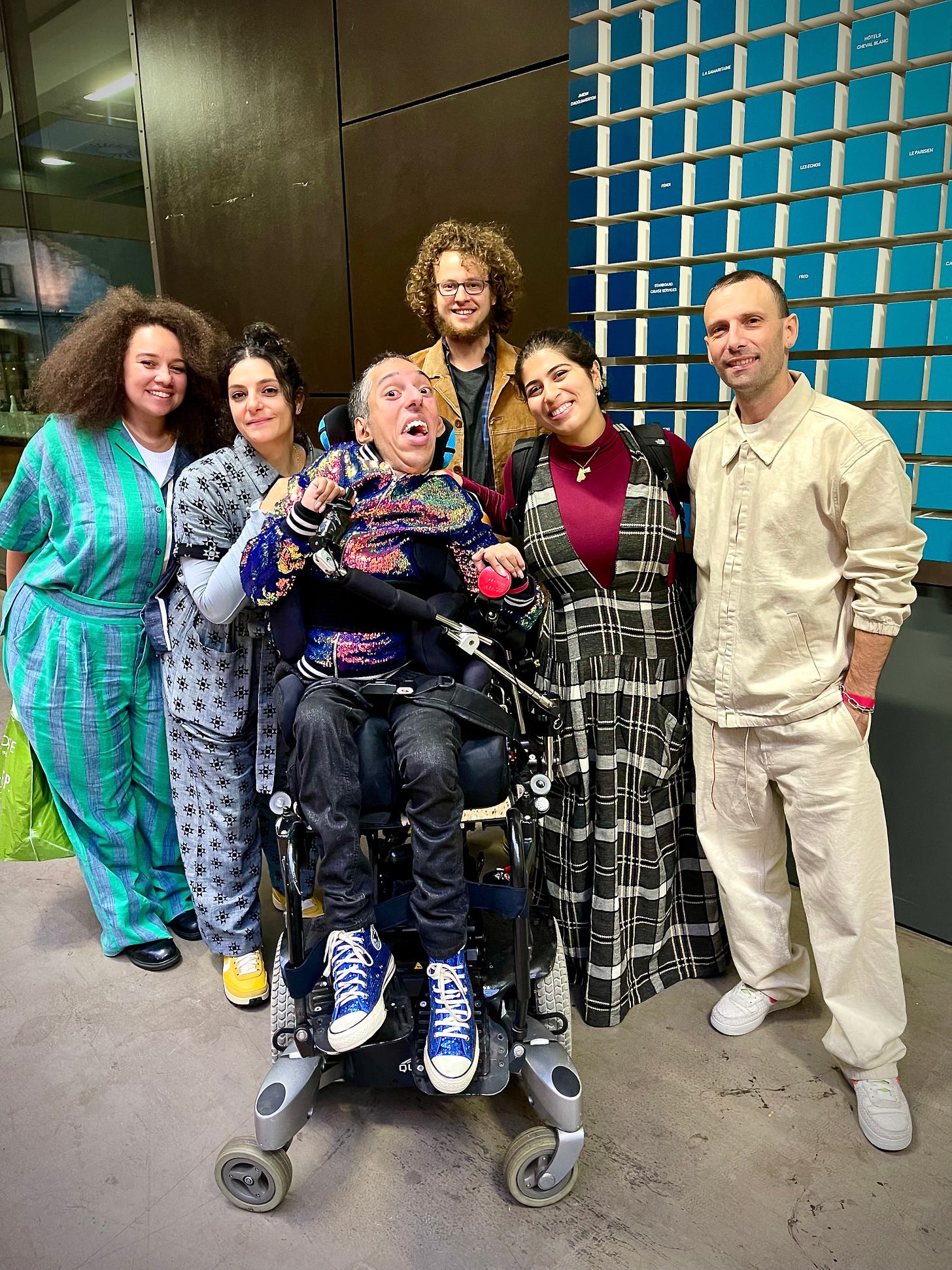 mage description 2: Bold, wild, fearless, existence. Group photo of Sulaiman (a wholeheartedly Disabled AF and British-Pakistani man with black buzzcut hair and features “white hair of wisdom”) sitting in his power wheelchair next to the team from The Slow Factory. All are smiling and joyful. END. 