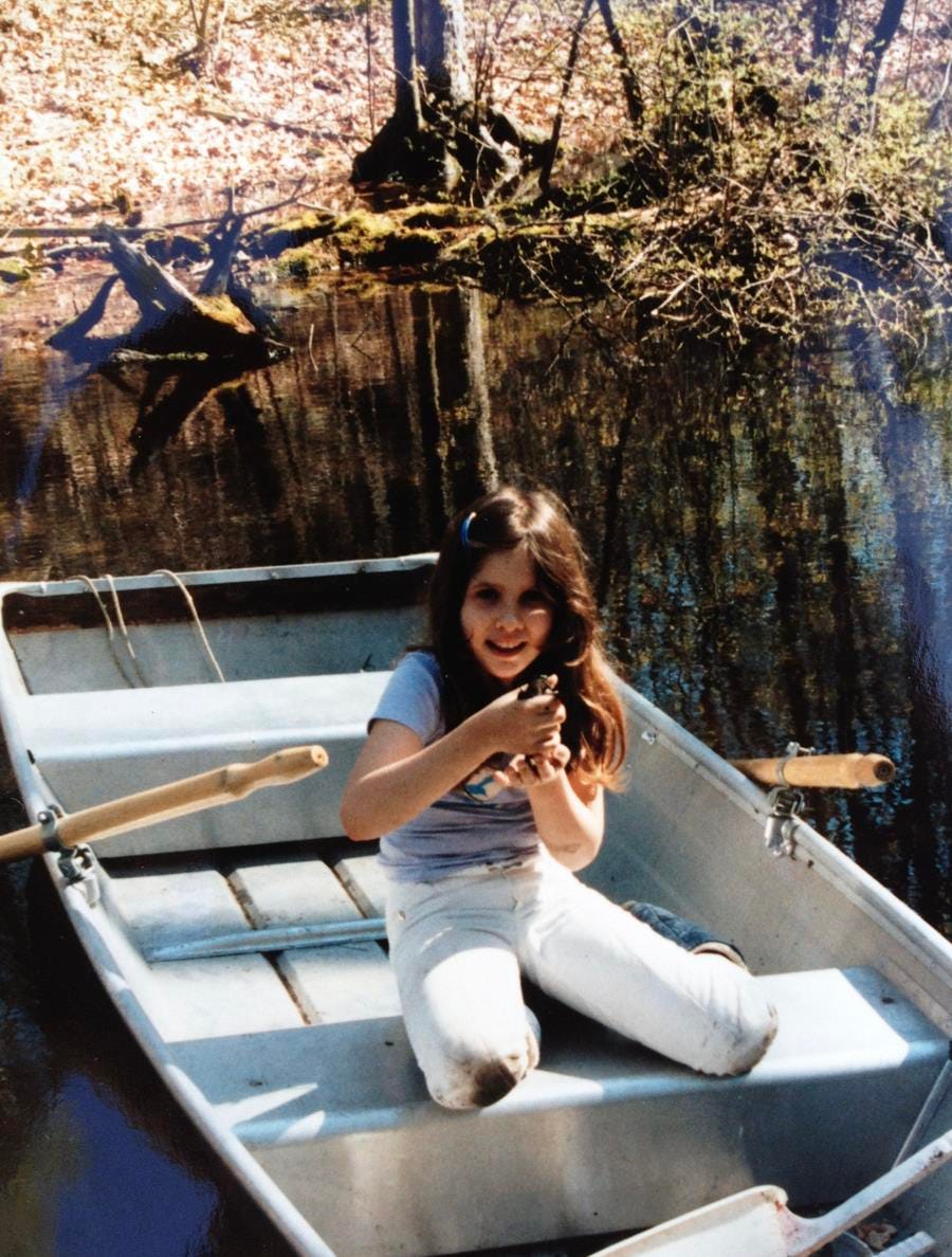 A young girl sits in a rowboat that is on floating on a small pond, holding a frog, the knees of her white jeans muddy.