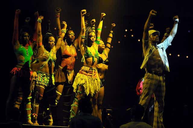 A stage-lit setting where a man with his shirt unbuttoned stands with both fists in the air. Behind him five women emulate his pose.