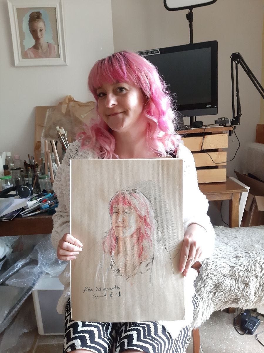Kim, smiling, with bright pink hair, holding a portrait of herself with closed eyes Kim, smiling, with bright pink hair, holding a portrait of herself with closed eyes