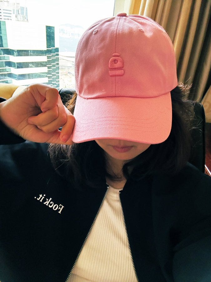 A person wearing a pink baseball cap with a small logo on the front. The individual is dressed in a black jacket with the text "i.rock" on it and a white top underneath. The person is sitting indoors near a window with a cityscape visible in the background.
