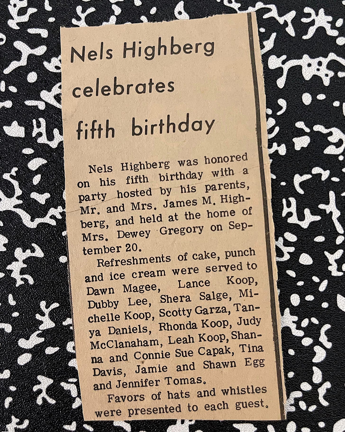 A yellowed newspaper clipping with the headline "Nels Highberg celebrates fifth birthday," announcing a small birthday party hosted by Nels's parents. The article lists the names of children who attended and describes refreshments like cake, punch, and ice cream. The clipping rests on a black-and-white speckled background resembling a classic composition notebook cover.