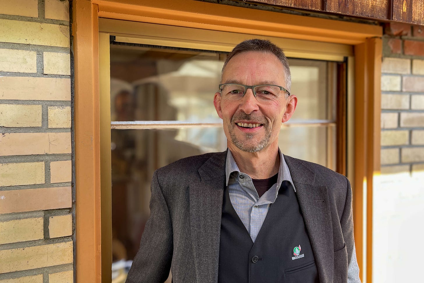 Urs Brändli, Präsident von Bio Suisse, steht mit kurzer Frisur, Brille und leichtem Bart lächelnd vor einer Backsteinwand und einem Fenster. Er trägt ein helles Hemd, eine dunkle Weste mit Bio-Suisse-Logo und ein graues Sakko.