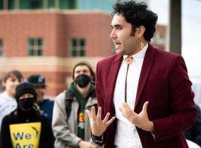 person in red blazer and bolo tie speaks to outdoor crowd