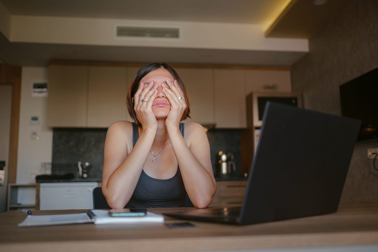 A woman in a tank top sits at a kitchen table with her face buried in her hands, looking distressed in front of an open laptop. A woman in a tank top sits at a kitchen table with her face buried in her hands, looking distressed in front of an open laptop.
