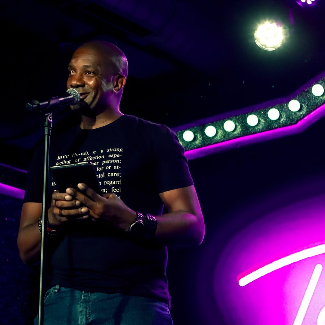 A close-up of Rick, a shaven headed, clean shaven black man speaking into a microphone. He is wearing a black t-shirt with white text on it. The text is a definition  of the word "love", he is holding a kindle tablet in both hands with interlaced fingers. The stage back drop is a string of white lights and accents of pink neon.