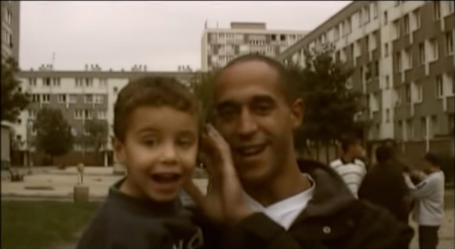 Screenshot extrait du clip du groupe de rap français 113, intitulé “Les Princes de la Ville” sorti en octobre 1999. Un plan rapproché sur un homme portant un enfant dans ses bras (probablement son fils) et souriants à la caméra. A l'arrière-plan, des bâtiments de banlieue parisienne.