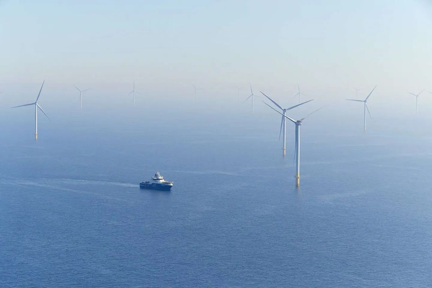 A small ship sails through an offshore wind farm. Several huge wind turbines are in the foreground, but the view of others stretches off to the misty horizon. A small ship sails through an offshore wind farm. Several huge wind turbines are in the foreground, but the view of others stretches off to the misty horizon.