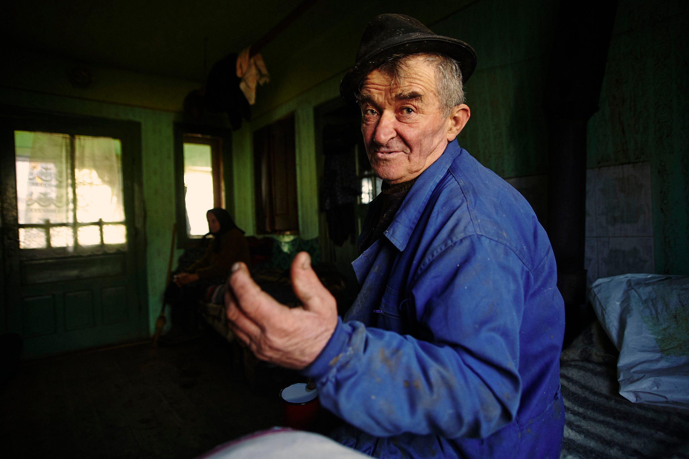 An older man in a blue jacket and black hat gestures with his hand while sitting in a dimly lit room. Another person is seated in the background near a door, with sunlight streaming through the windows.
