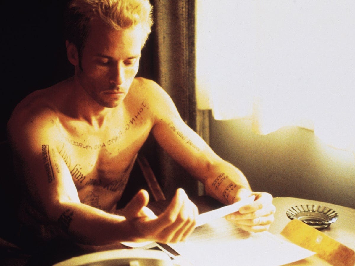 Man in his early 30s sitting at the table, reading what looks like a note, in a dimly lit hotel room. His body is covered with tattoos. He looks serious, focused.