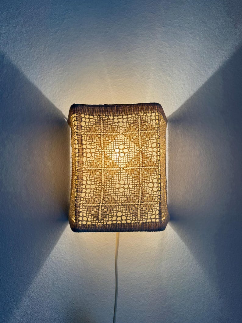 May include: A square wall sconce with a woven, openwork design. The shade is a light tan color, and the frame is a darker brown. The sconce is lit from within, casting a warm glow and creating a pattern on the wall. May include: A square wall sconce with a woven, openwork design. The shade is a light tan color, and the frame is a darker brown. The sconce is lit from within, casting a warm glow and creating a pattern on the wall.