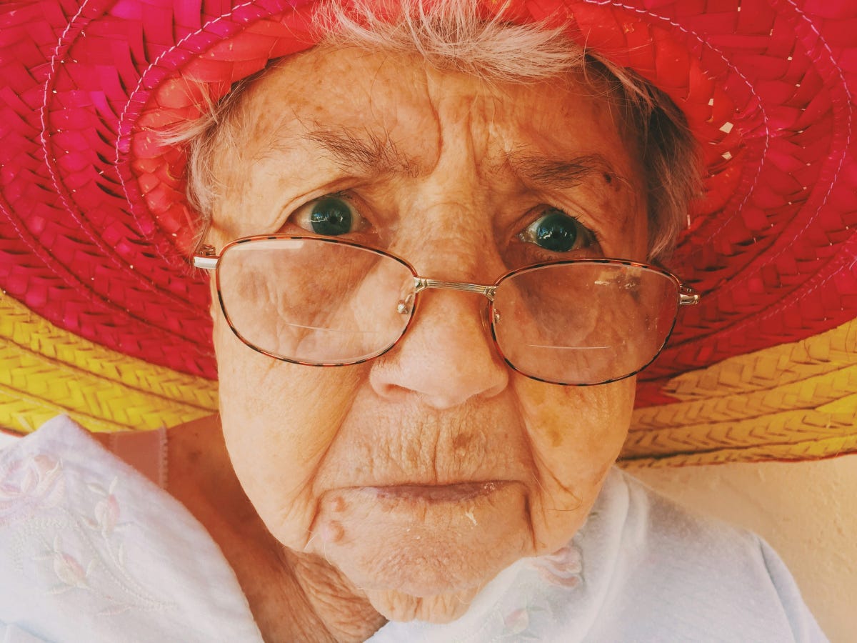 Elderly woman with red and yellow straw hat and white blouse, staring with blue eyes over tortoise-framed eyeglasses