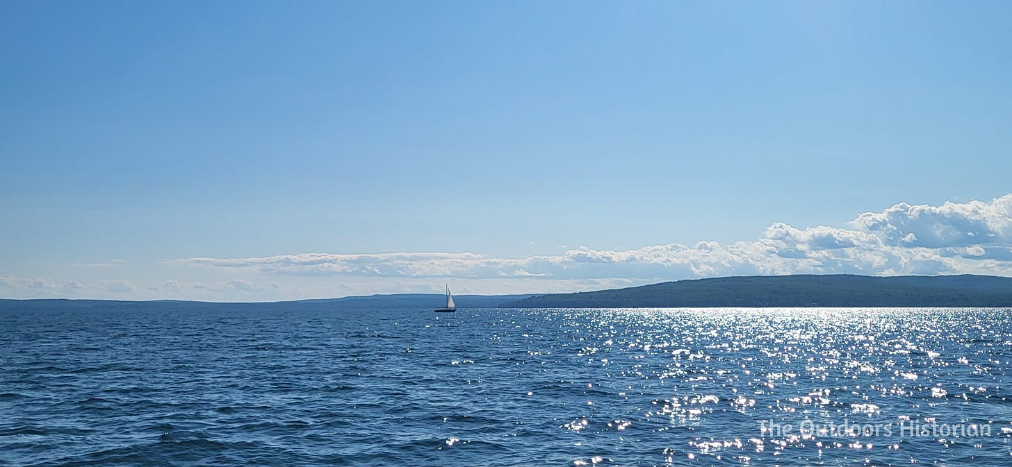 An image showing a blue sky, a blue lake, and forested hills in the background.