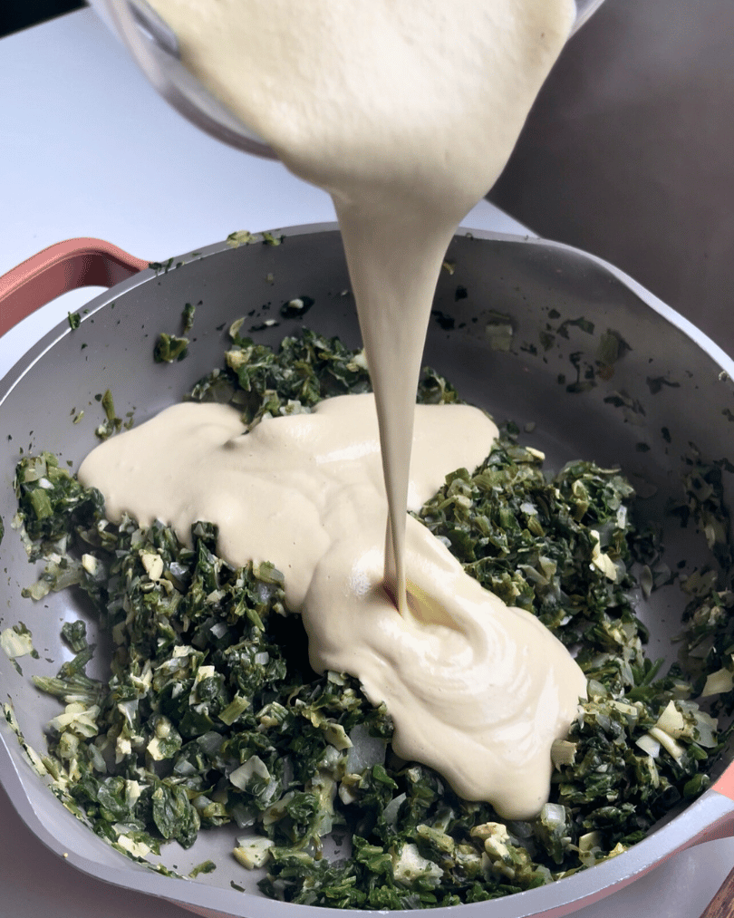 cooked spinach, artichoke, and onion in a pan with a creamy sauce being poured into it