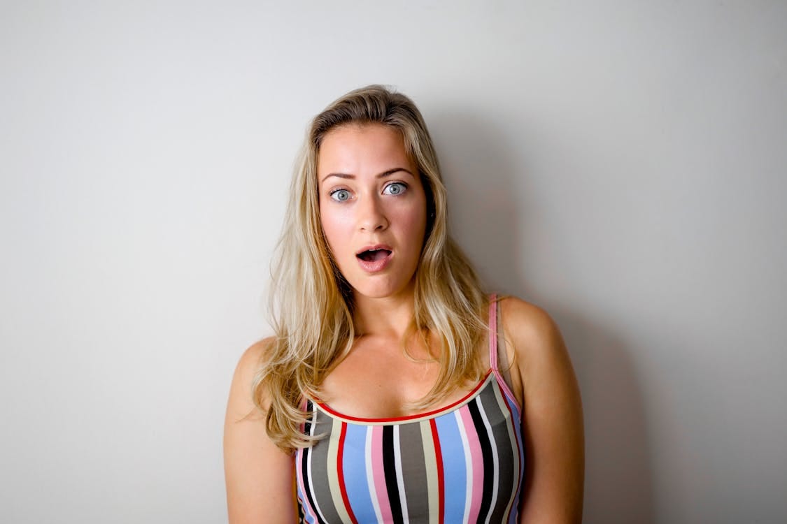 Free A young woman with a shocked expression stands against a plain white wall. Stock Photo