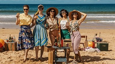 A group of 5 women having fun on a sunny beach. Dressed in vintage clothes from the 1950s, they have drinks, picnic baskets, a beach ball, radio, cool boxes and are smiling in their sunglasses
