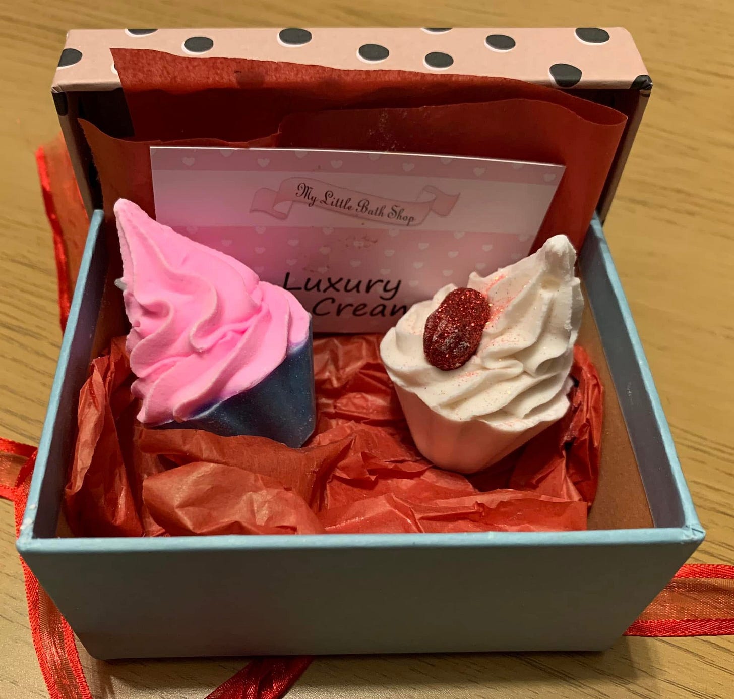 Image of two bath bombs that look like cupcakes in an open box against a background of red tissue paper