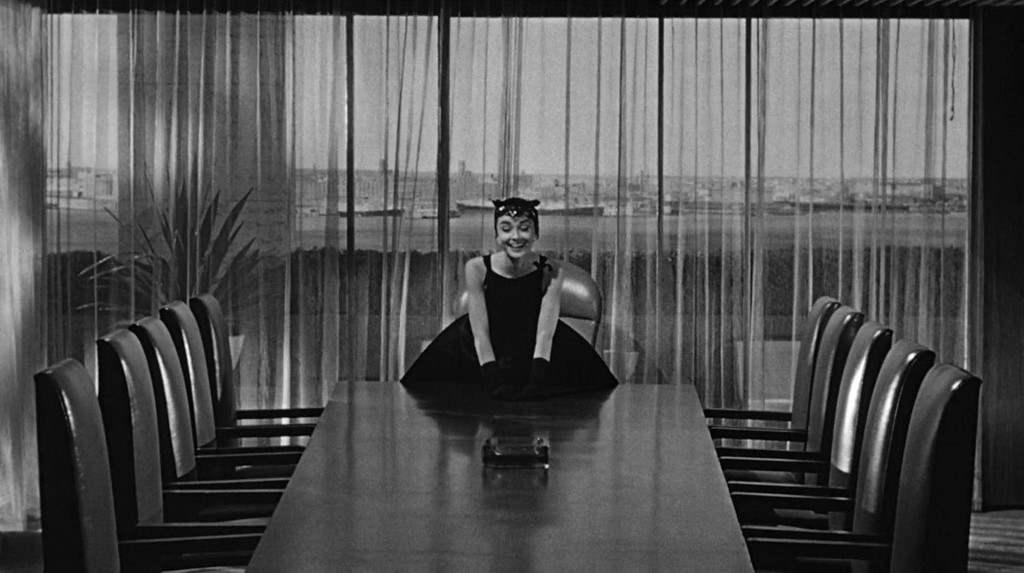 Hepburn in a black dress and a fancy skull cap sitting at the end of a boardroom table with a harbor in the background through floor length windows. She's smiling and has her hands on the tableAubrey Hepburn centered; wearing a black dress with gloves and bare arms; she’s standing at the end of a boardroom table laughing and leaning on the table. Full length window behind her with curtains; a harbor is visible beyond them. Hepburn in a black dress and a fancy skull cap sitting at the end of a boardroom table with a harbor in the background through floor length windows. She's smiling and has her hands on the tableAubrey Hepburn centered; wearing a black dress with gloves and bare arms; she’s standing at the end of a boardroom table laughing and leaning on the table. Full length window behind her with curtains; a harbor is visible beyond them.