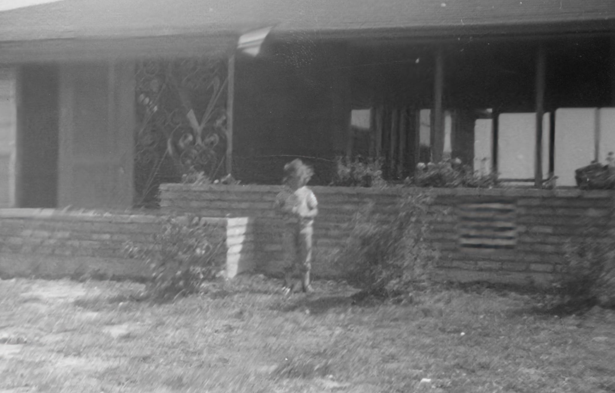 A blurred black-and-white photo of a child standing by a brick wall in front of a house with a partially visible porch.