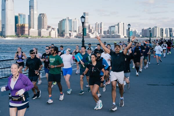 A large group of runners run on a path alongside the water. Tall buildings in Jersey are seen across the river in the background. A large group of runners run on a path alongside the water. Tall buildings in Jersey are seen across the river in the background.