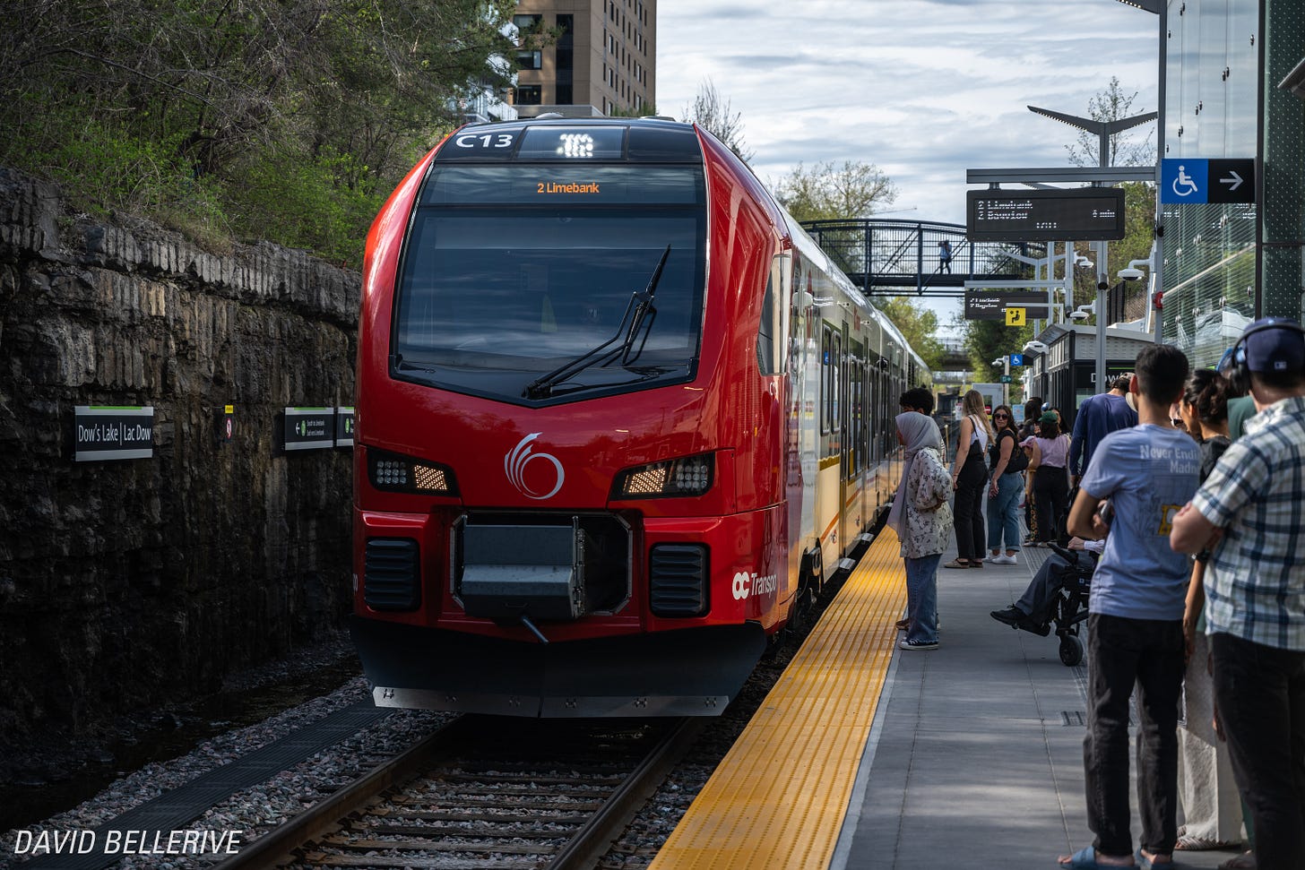 Passengers waiting on the Dow’s Lake O-Train platform as a red Stadler FLIRT train arrives on a sunny spring day.