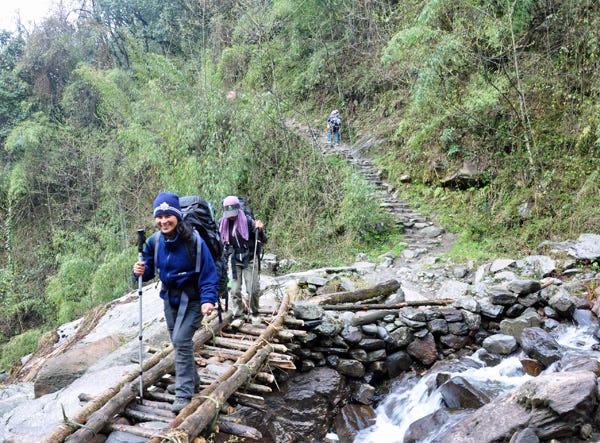 Two young Nepali women wearing hiking gear crossing a wooden bridge in the mountains. 