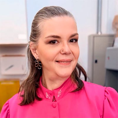 Fotografia de uma mulher adulta em um ambiente que parece ser um laboratório ou consultório odontológico. Ela está em pé, de frente para a câmera, sorrindo levemente. Tem cabelos castanhos presos para trás e usa uma roupa profissional rosa de mangas compridas, com o nome “Profa. Fernanda Panzeri” bordado na parte da frente. Ao fundo, há equipamentos e móveis de laboratório. Fotografia de uma mulher adulta em um ambiente que parece ser um laboratório ou consultório odontológico. Ela está em pé, de frente para a câmera, sorrindo levemente. Tem cabelos castanhos presos para trás e usa uma roupa profissional rosa de mangas compridas, com o nome “Profa. Fernanda Panzeri” bordado na parte da frente. Ao fundo, há equipamentos e móveis de laboratório.